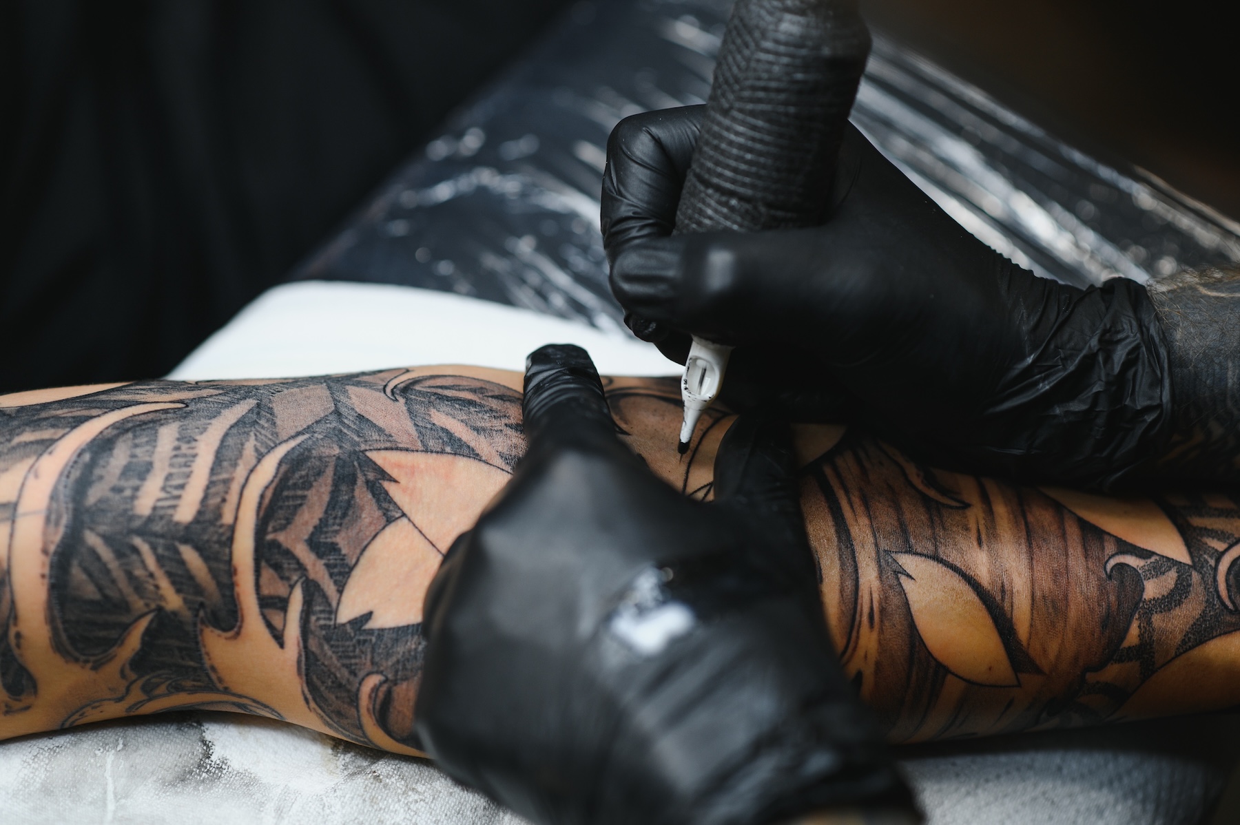 Cropped close up of a bearded tattoo artist working at his studio tattooing sleeve on the arm of his male client. Man getting tattooed by professional tattooist