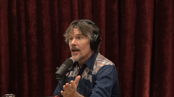 Ethan Hawke sitting in front of a red curtain and gesturing with his hands while he speaks into a microphone.