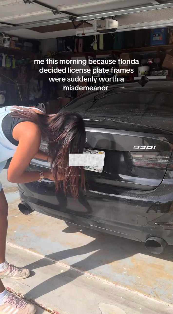 A woman bent down to remove her license plate frame from her car, her face obscured by her hair.