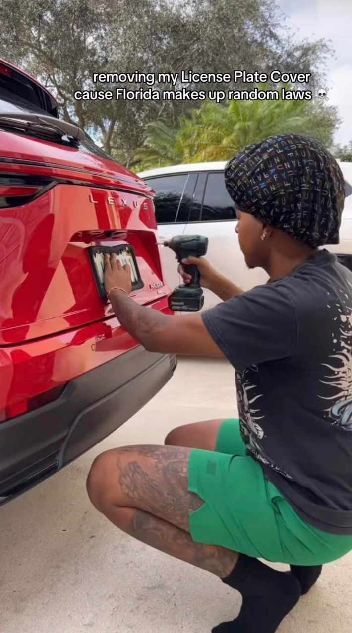 A man crouched down behind his car, carefully removing the license plate frame with a drill.
