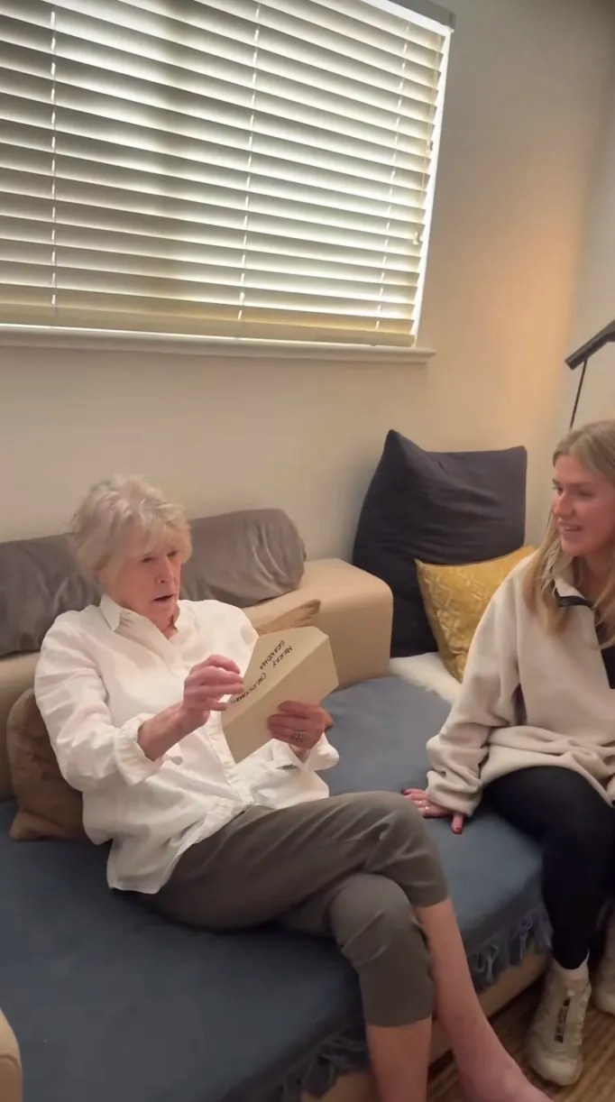 A blonde woman sitting next to her grandmother on a couch while the older woman opens an envelope.