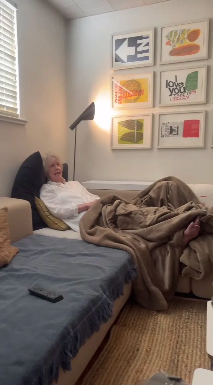 An older woman lying cozy on her couch with a blanket draped over her lap.