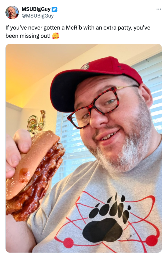 Tweet that reads, "If you’ve never gotten a McRib with an extra patty, you’ve been missing out! 🥰" with a photo of a smiling man in a red baseball cap and grey t-shirt holding up a double McRib sandwich.
