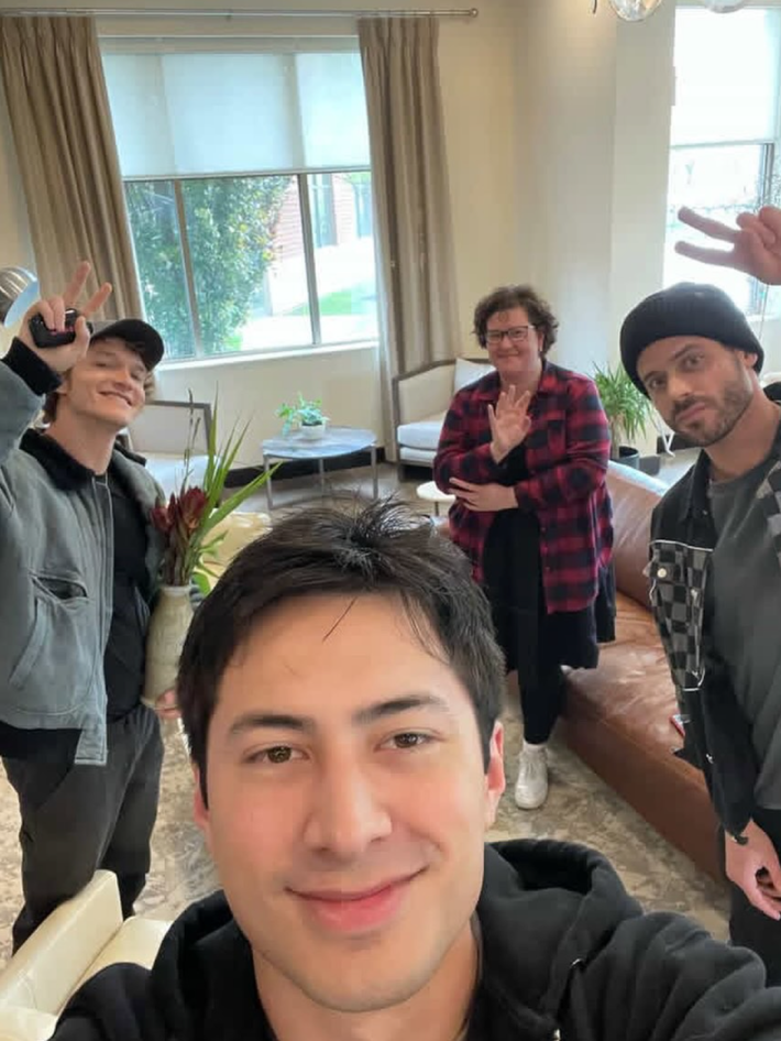 A group selfie with actors Hudson Williams, Connor Storrie, and François Arnaud and romance author Rachel Reid in the background. Connor and François are giving peace signs to the camera.