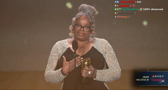 An older Black woman wearing a sparkly silver and black dress with her silver hair in an updo giving a thank you speech while holding her gold statue award.