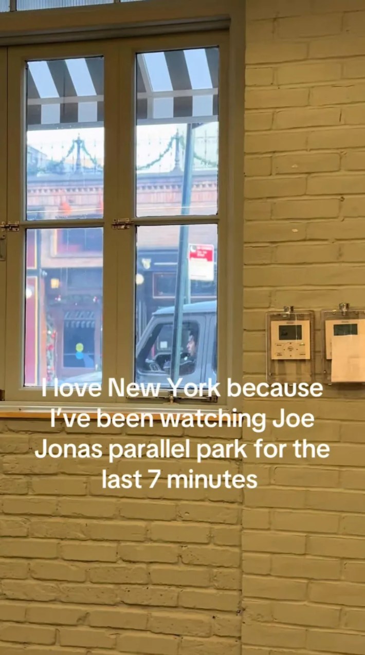 View from the inside of a white brick building looking out the window to Joe Jonas trying to park his car.