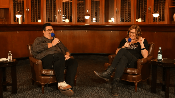 Lilly Wachowski and Caleb Hearon sitting in brown armchairs and speaking into handheld mics. Both are laughing with their legs crossed in front of them.