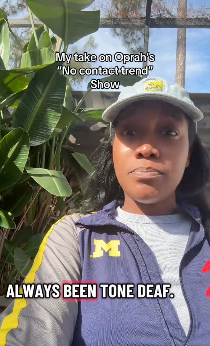 A woman talking to her camera, sitting in front of a large plant while she explains that Oprah has "always been tone deaf."