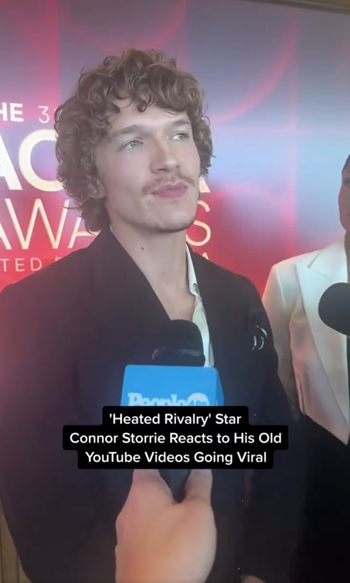 Connor Storrie on the red carpet at the Actor Awards nominations announcement.  He is speaking into a mic. Text overlay reads, "Heated Rivalry star Connor Storrie reacts to his old YouTube videos going viral."