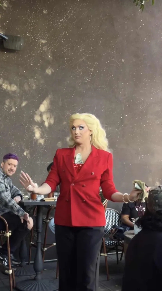 A drag queen dressed as Erika Kirk staring down the crowd. She is wearing a bright red blazer and ice blue contacts and has a dollar bill stuffed in her bra, holding more money in her placating raised hands.