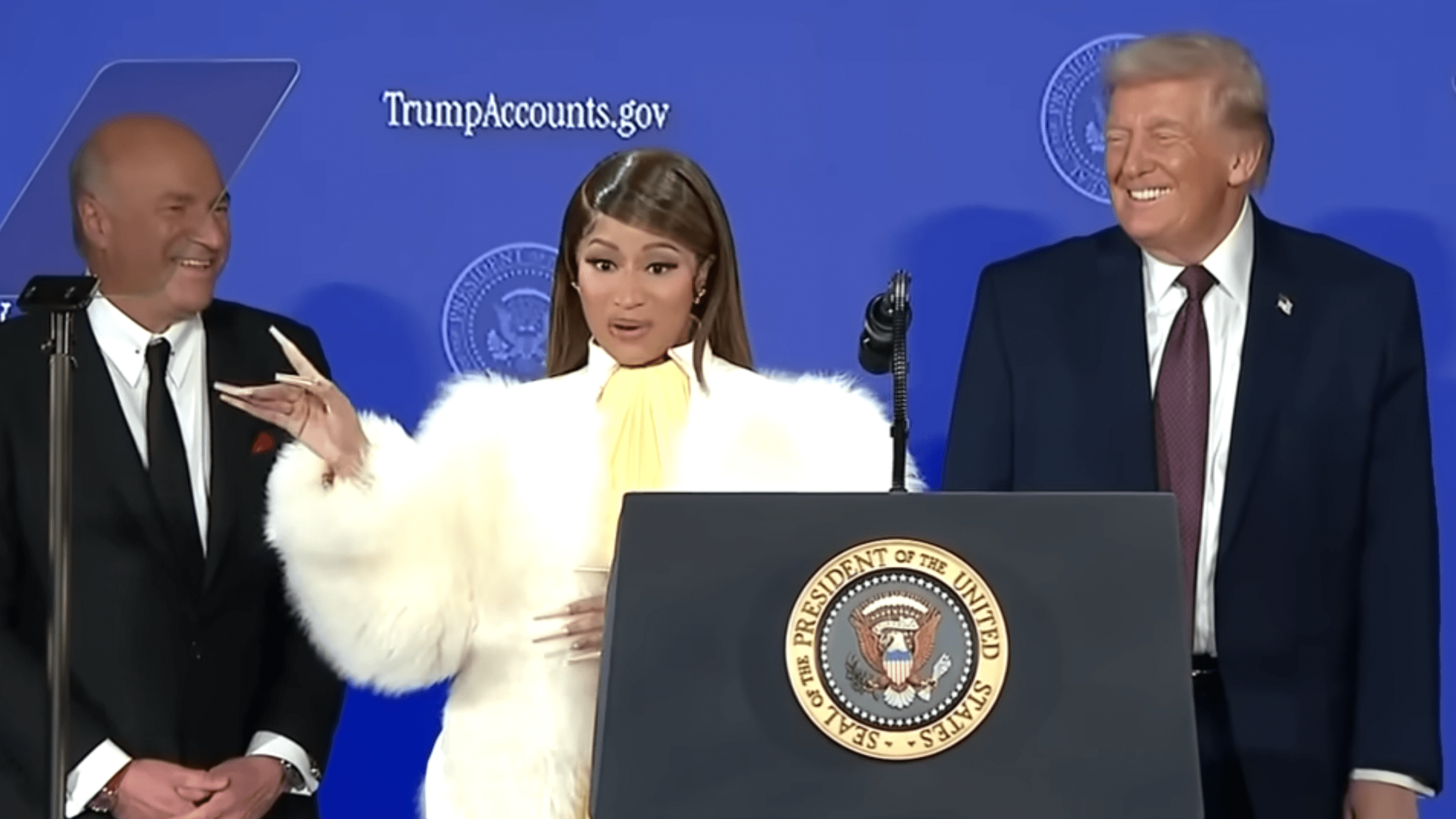 Nicki Minaj on stage with Donald Trump and Kevin O'Leary standing in front of a podium