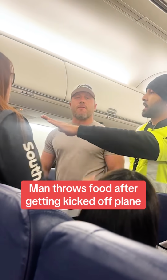 Angry male passenger glaring down the camera, a pizza box in hand like he's trying to figure out how to throw it. There is a flight attendant and security surrounding him to get him off the plane. Text overlay reads, "Man throws food after getting kicked off plane."