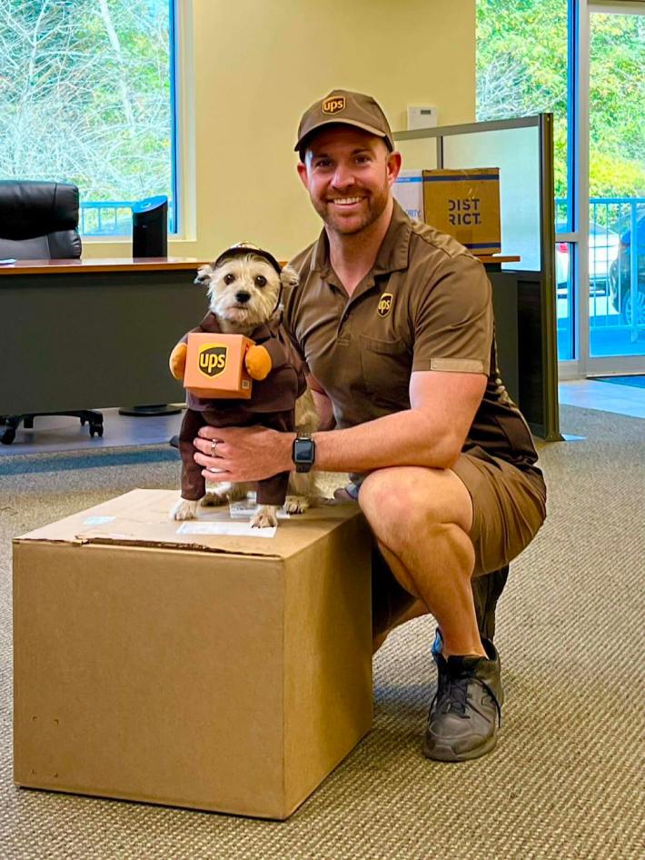 A UPS driver crouching next to a tiny white dog in a UPS driver costume standing on a big cardboard box and "holding" a box of its own.
