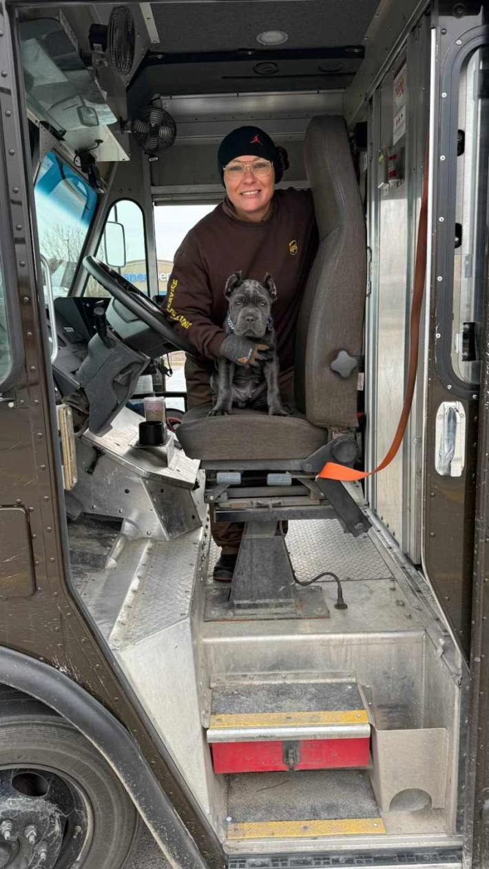 A bundled-up UPS driver standing in the cab of her truck, her arm around a small black dog sitting on her driver seat.