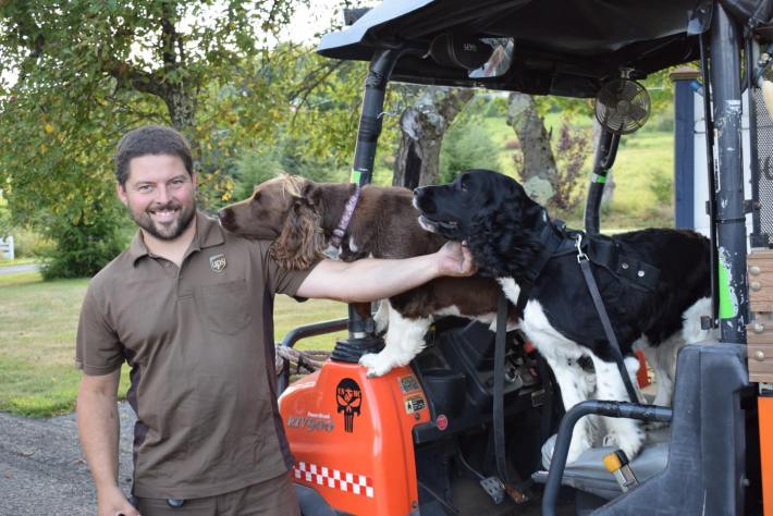 A UPS driver standing next to a golf cart with two floppy-eared dogs staring lovingly at him from the seat. One with her chin on his shoulder.
