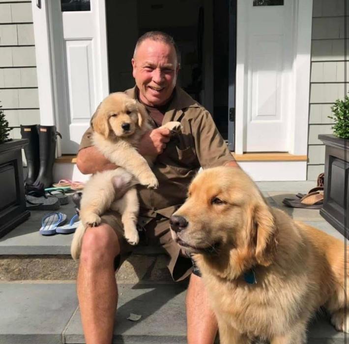 A UPS driver sitting on a porch, a big golden retriever sitting next to him and a puppy in his lap.