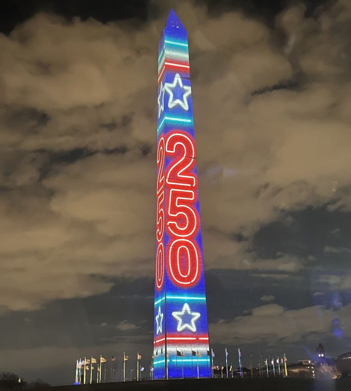 The Washington Monument under a cloudy night sky with bright red, white, and blue stars and "250" projected on its sides.