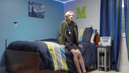 Um menino sentado em uma cama usando um colar branco com um ponto branco, o colar do amigo.