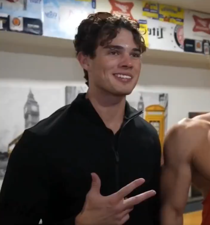 Clavicular wearing a black button up shirt and giving a three-fingered peace sign as he poses for a photo next to a muscular man.
