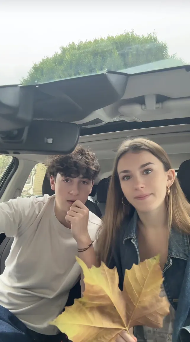 A young couple sitting in a car, the woman is holding up a massive maple leaf as the man looks confused at the camera.