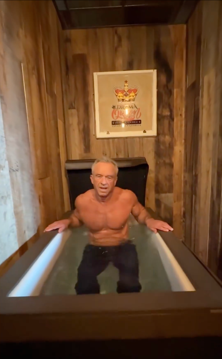 RFK Jr sitting in a cold plunge pool shirtless and in full jeans. A poster on the wall behind him reads, "Drama Queen."