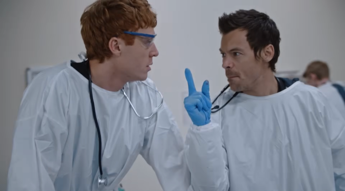 Two men in fake PPE, the one with blue gloves gesturing with one hand while giving an order for fake medicine.