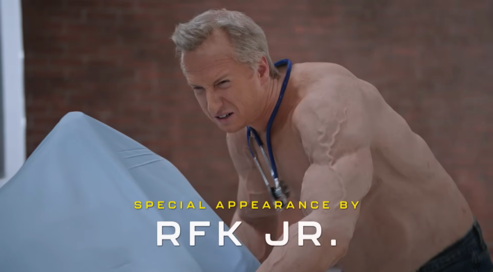 A man with grey hair and a fake muscle suit and no shirt parodying RFK Jr. squinting as he pushes a gurney into a fake hospital.
