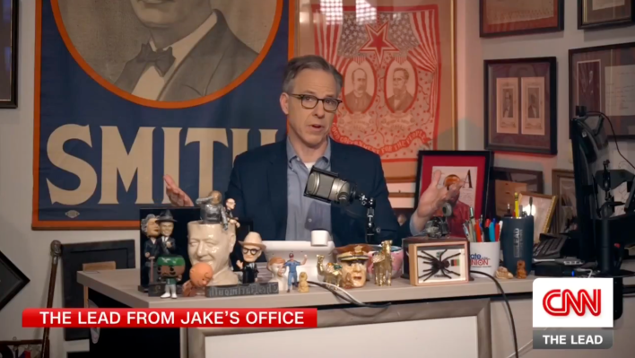 Jake Tapper sitting behind a messy desk filled with knickknacks including bobble heads and a spider in a glass case. The walls are covered with campaign posters with almost no white space.