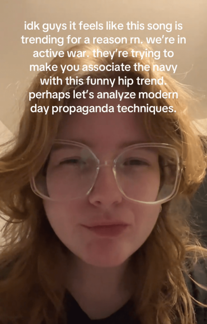 Redheaded person in large framed glasses squinting their eyes at the camera. Text overlay reads, "idk guys it feels like this song is trending for a reason rn. we're in active war they're trying to make you associate the navy with this funny hip trend. perhaps let's analyze modern day propaganda techniques."