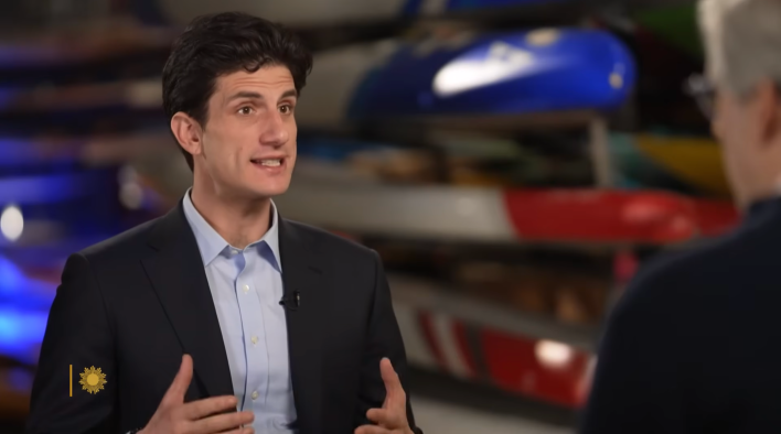 Jack Schlossberg in an interview wearing a suit and no tie, gesturing with his hands as he speaks.