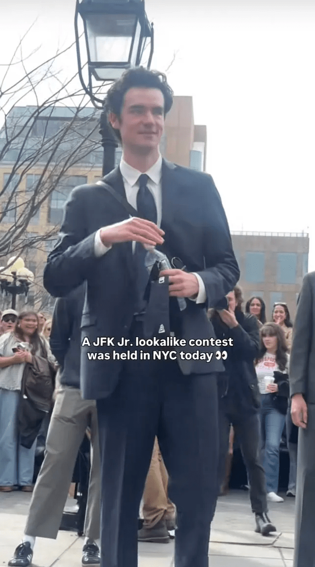Man in a business suit holding a water bottle and baseball cap standing in front of a crowd of people.