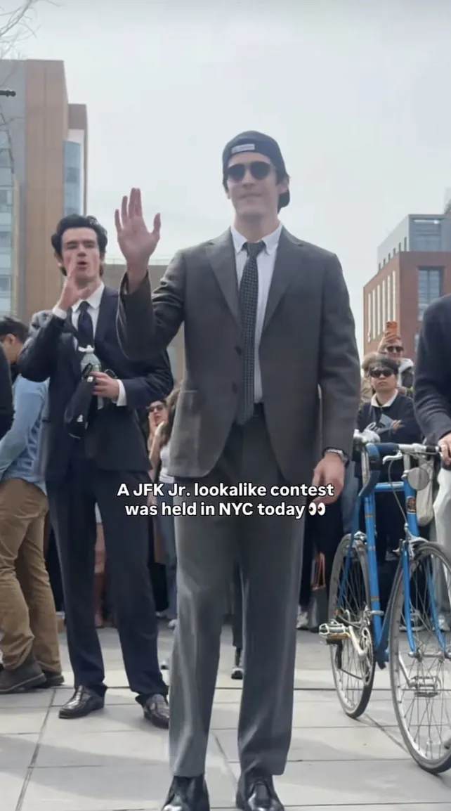 Man in a business suit wearing sunglasses and a backwards baseball cap, hand raised to the crowd of people around him.