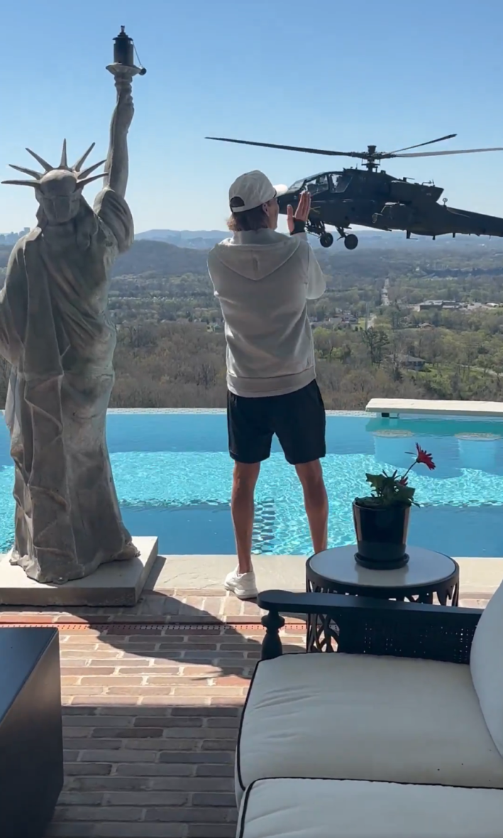 Kid Rock standing behind his pool with a miniature version of the Statue of Liberty applauding an Apache helicopter hovering by his Nashville home.