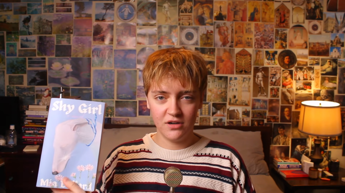 YouTube video of a short-haired person sitting in front of a wall filled with unframed photographs. They are holding up a copy of Shy Girl by Mia Ballard and look displeased.
