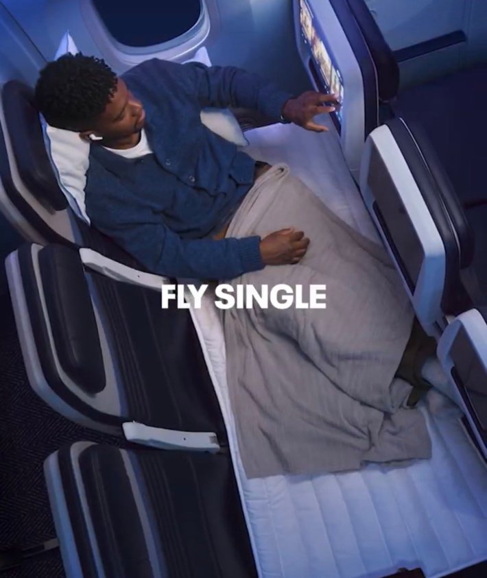 A man stretched out on a row of three seats that has been folded out into a bed, clicking through the seat back movie menu. Text overlay reads, "Fly single."