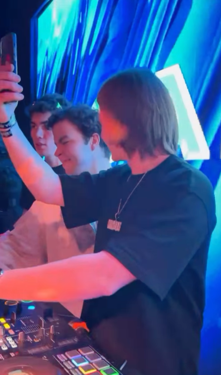 Three men standing behind a night club DJ booth with a full LED screen behind them.